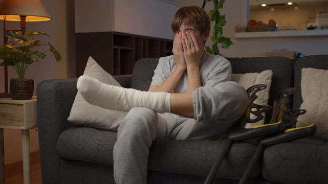 Young male patient recovering from broken leg sits at home with plaster cast, massaging injured limb while adapting to rehabilitation, limited movement and healing process.