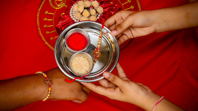 Top-down view of a sister's hands holding a traditional puja thali containing roli, chawal, and a rakhi, next to her brother's wrist during the Indian festival of Raksha Bandhan