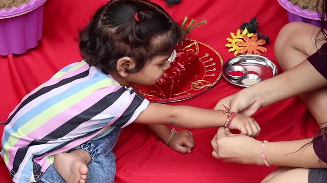 Older sister tying a traditional rakhi thread on her cute younger sister's wrist during Raksha Bandhan festival celebrations on a vibrant red background.
