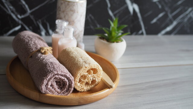Luxury eco-friendly bathroom setup with a bamboo toothbrush, natural loofah, and rolled pink towel on a wooden tray, paired with a white marble ceramic soap dispenser. Side view on a light gray wood 