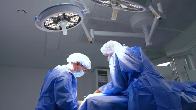Cooperating male surgeons stand at the operating table under the bright round lamps. Low angle view at the doctors performing foot surgery.