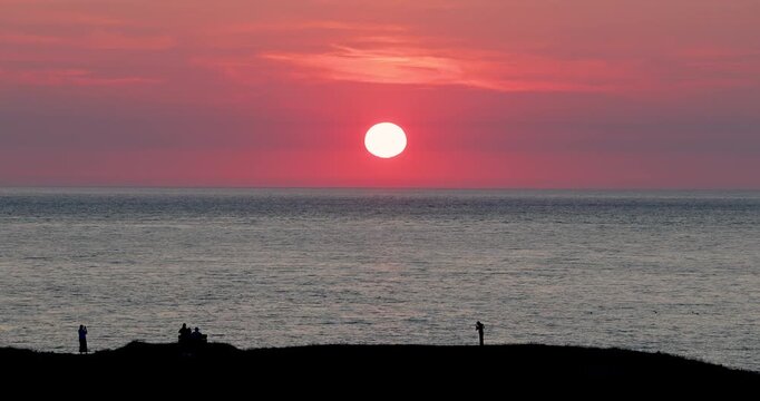 beautiful midsummer sunset setting over the sea with silhouetted people in the foreground admiring the stunning view as the sun sets in a dramatic red evening sky over the ocean 4K landscape video