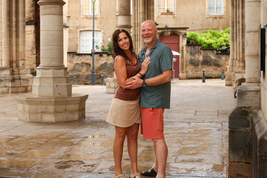 Smiling couple standing close together in a stone courtyard after a rain shower in Beaune, France, surrounded by historic architecture and soft natural light.