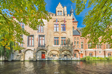 Fototapeta premium Bruges town in Belgium, Europe. Historic European Canal View with Ancient Architecture and Lush Green Foliage on a Bright Sunny Day, Reflecting Tranquil Waters Below