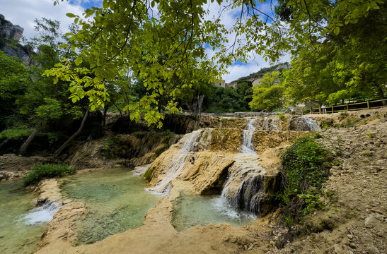peaceful waterfall cascading over rocks in lush green forest setting with trees and pathway ground-level perspective natural landscape. Risco los camellos, Cascada de Orbaneja del Castillo