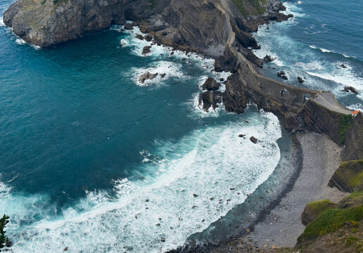 cliffside pathway with crashing waves on rocky coastal cliffs, aerial view of natural scenery with blue and white ocean waves and concrete and gravel materials.