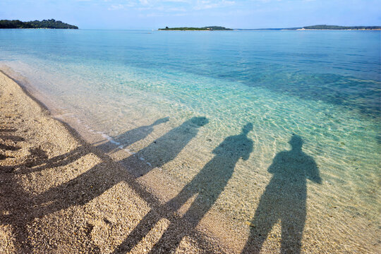 Four elongated human shadows stretch across a pebble beach beside transparent turquoise water, forming a creative summer travel scene with distant islands and calm coastal atmosphere.