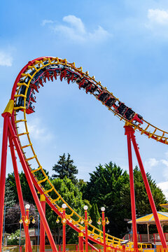 Roller coaster riders speed along a dramatic curved track beneath a blue sky, creating a vivid amusement park scene filled with motion, adrenaline and summer excitement.