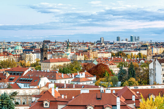Autumn view of Prague with red rooftops, colorful trees, and warm city lights under a partly cloudy sky, capturing the charm of the historic European capital.