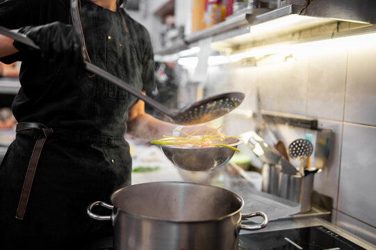 A chef wearing a black apron and gloves uses a slotted spoon to strain food over a steaming pot in a commercial kitchen