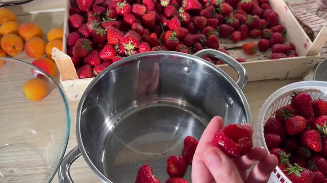 Hand holding fresh strawberry over stainless pot on kitchen counter, surrounded by baskets of berries and apricots, bright home canning prep mood
