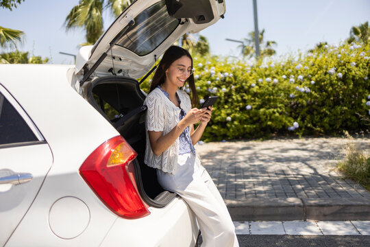 Young woman texting by open car trunk in sunny parking lot
