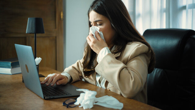 Young Hispanic businesswoman working in her office while suffering from severe nasal congestion and feeling unwell, trying to stay productive despite her physical discomfort.