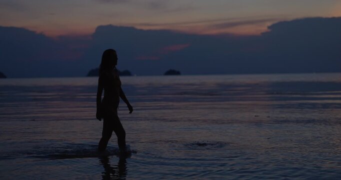 Woman walks across shallow shoreline in dark silhouette under blue dusk sky on holiday. Female swimmer keeps steady pace moving past calm ocean during twilight