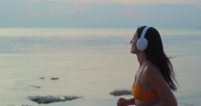 Middle Eastern woman runs in orange bikini with white headphones near calm sea at dusk. Runner keeps arms bent maintaining steady pace during coastal workout