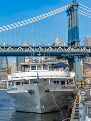Fototapeta premium New York, USA, 4 Jule 2026 : Grande Mariner ship bow docked under Manhattan Bridge in New York. Close up front view of white cruise boat moored by pier with blue suspension bridge tower behind.