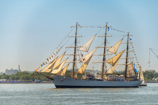 Tall ship USCGC Eagle sails through New York Harbor. Three masted barque displays signal flags and cream sails over blue water with trees and port cranes behind.