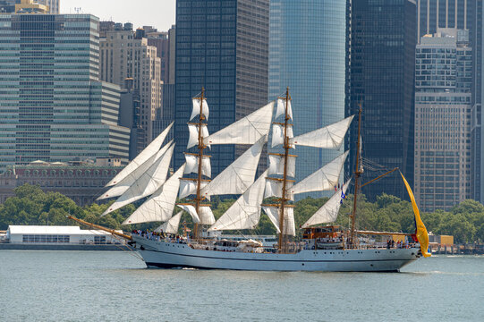 BAE Guayas sails before Lower Manhattan on Independence Day. Ecuadorian Navy barque carries white sails and a yellow stern flag against dense waterfront office towers.