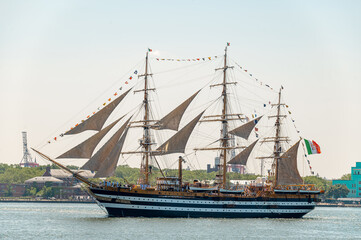 Obraz premium Amerigo Vespucci sails through New York Harbor on July 4. Italian Navy training ship carries dark sails, signal flags and an Italian flag over blue water.