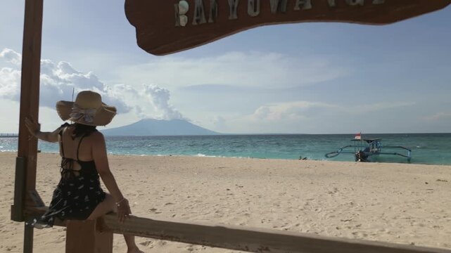 Low angle aerial view Girl sitting on bench at Tabuhan Island beach looking at the sea view with volcano in the distance and small Jukung boat and blue sky in the background in East Java, Indonesia
