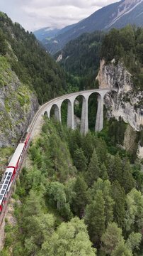 Portrait aerial drone footage of the iconic Landwasser Viaduct, following a train, captured from the air in the dramatic alpine landscape