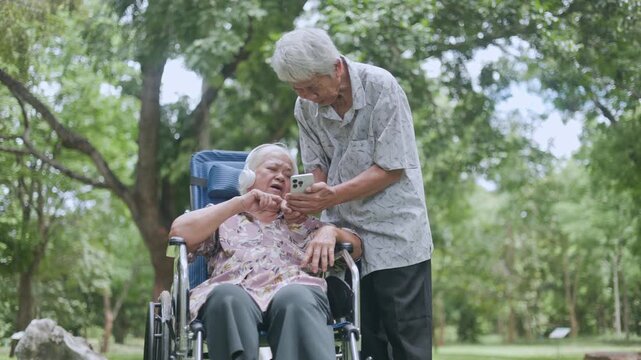 Caring young man helps his old handicapped mother adjust her white headphones while using a smartphone together, showing deep family love and dedicated caregiving lifestyle.