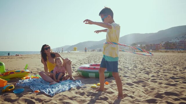 Young boy hula hooping on the sandy beach while his mother and toddler sister watch him, all enjoying a sunny summer day. Carefree family having fun together during their seaside vacation