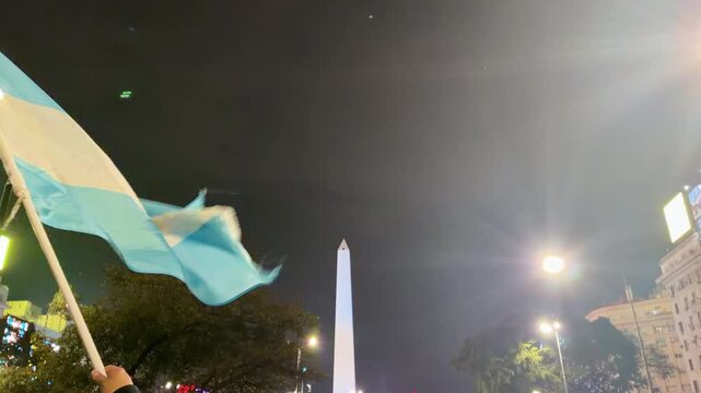 BUENOS AIRES, ARGENTINA - JULY 2026: Close up of a person waving Argentina national flag with the iconic Obelisco monument in the background during world cup celebrations.