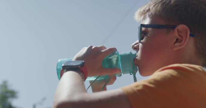On a sunny, hot day, a boy drinks water from a portable personal bottle to quench his thirst.