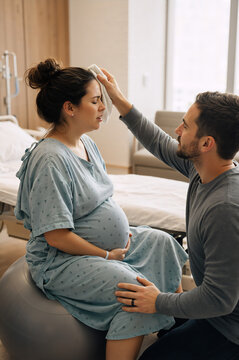 Husband supporting pregnant woman during labor in hospital room. Partner assisting wife with wet towel during contraction. Expectant couple preparing for childbirth experience indoors.