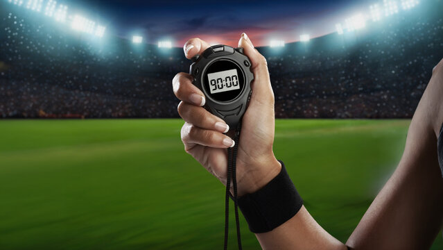 Close up of a female hand with a black wristband holding a digital stopwatch showing ninety minutes with a large football stadium blurred background, concept of match full time