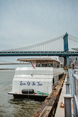 Fototapeta premium New York, USA, 2 May 2026: White cruise ship docked on East River near Manhattan Bridge. Back side of tour boat at the pier with Manhattan Bridge and Brooklyn Bridge visible in distance.