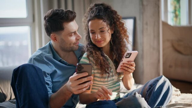 Couple laughing sharing phones home bedroom. Man and woman comparing smartphones
