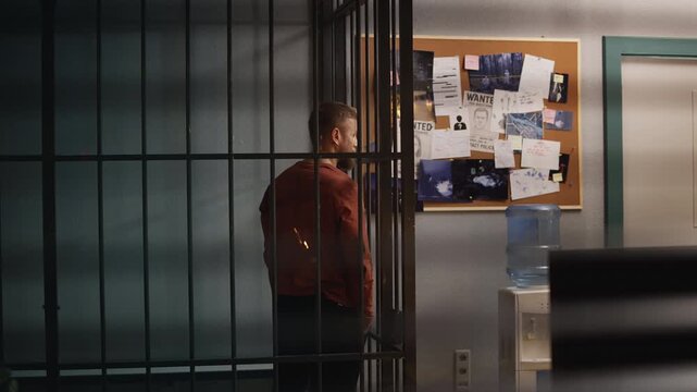 Young male suspect standing behind police cell bars, awaiting trial