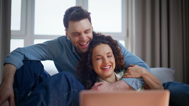 Couple embracing sofa watching pink laptop. Man woman laughing video call home.