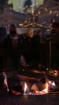 Hands Warming by Fire at Christmas Market in Winter Evening with People Walking in Background