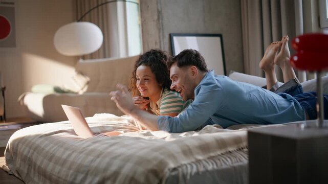 Laughing couple watching laptop lying bed. Happy pair relaxing home weekend.