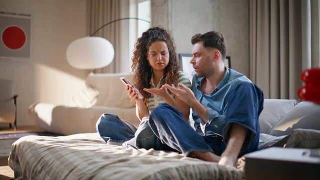 Worried couple checking phone together home discussing bad news sitting on bed