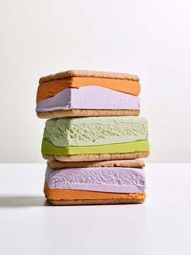 Stack of three colorful ice cream sandwiches featuring orange, purple, green layers with cookie wafers, placed on a white surface against a light background