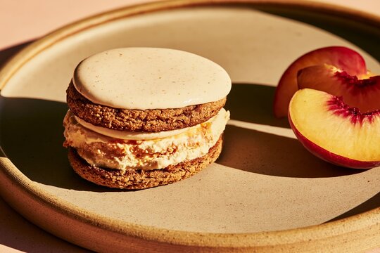 Gourmet ice cream sandwich with brown cookie layers and creamy filling, accompanied by a fresh peach slice on a beige plate against a soft background