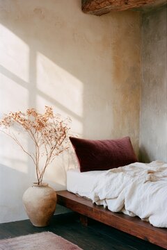 Wabi-sabi bedroom corner featuring a wooden bed with a white blanket, a burgundy pillow, and a ceramic vase with dried flowers against a textured plaster wall