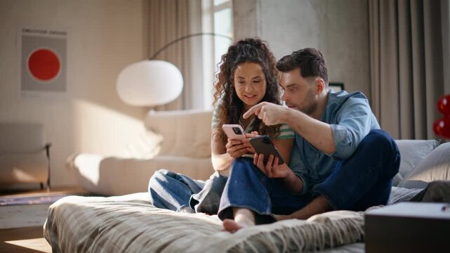 Couple comparing two phones sitting cozy bed. Man pointing screen smiling home.