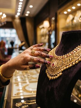 Woman's henna decorated hand touching intricate gold necklace displayed on velvet tray in luxury jewelry store setting.