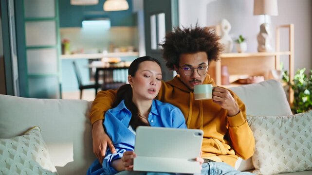 Multiethnic couple browsing tablet on sofa. Boyfriend drinking coffee hugging