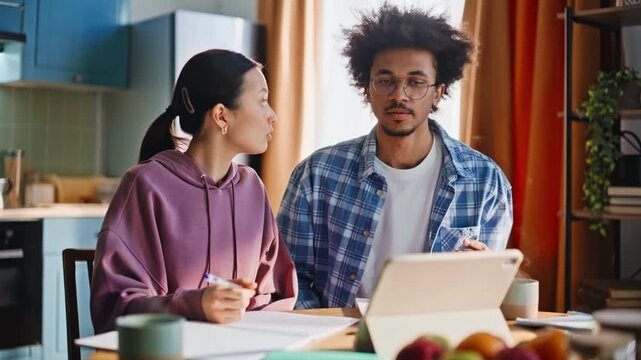 Couple studying together home kitchen. Multiethnic students taking notes tablet.
