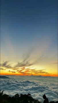 Vibrant sunrise colors paint the sky above a sea of soft clouds. A solitary figure and trees are silhouetted, witnessing this majestic, ethereal mountain dawn.