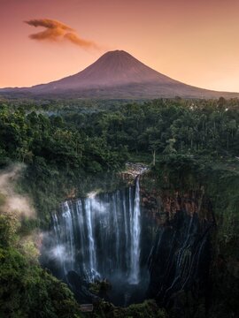 Majestic Tumpak Sewu waterfall at dawn, featuring the active eruption of Mount Semeru volcano in East Java, Indonesia. Epic jungle landscape with mist and a large ash cloud
