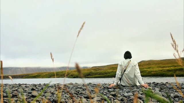 A lone figure sits on a rocky shore, gazing across a tranquil river towards distant, mist-shrouded mountains under an expansive, cloudy sky. Pure natural serenity.