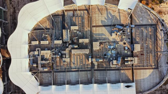 The scene shows a construction site with a complex layout and multiple structures. Workers and machinery are scattered across the space under a large roof.