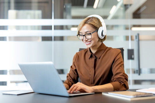 Smiling professional businesswoman engaging in remote work, wearing headphones and eyeglasses while typing on a laptop at a modern office desk, representing productivity and flexible working